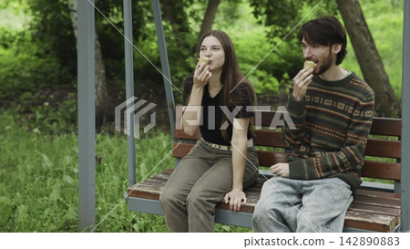 Two young adults sharing ice cream on wooden swing bench in park beneath soft overcast daylight, exchanging friendly looks with relaxed posture and casual outdoor mood Two young adults sharing ice cream on wooden swing bench in park beneath soft overcast daylight, exchanging friendly looks with relaxed posture and casual outdoor mood 142890883