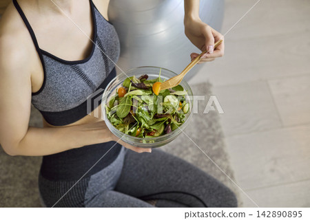 Fit woman in sportswear eating fresh salad at home after workout session Fit woman in sportswear eating fresh salad at home after workout session 142890895