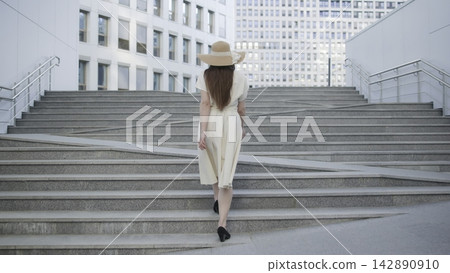 Woman in light dress and wide hat walking up broad city steps between modern buildings Woman in light dress and wide hat walking up broad city steps between modern buildings 142890910