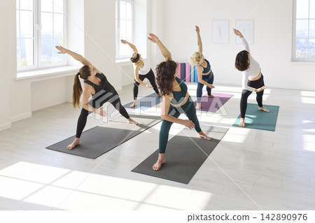 Group yoga class practicing side angle pose in bright minimalist studio interior Group yoga class practicing side angle pose in bright minimalist studio interior 142890976