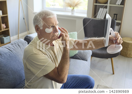 Senior man applying under eye patches with mirror at home, skincare routine Senior man applying under eye patches with mirror at home, skincare routine 142890989