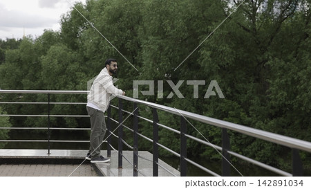 Man leaning on stainless steel railing at riverside viewing platform, gazing toward tree lined river beside park greenery beneath soft overcast daylight, with man, leaning, railing, beard detail Man leaning on stainless steel railing at riverside viewing platform, gazing toward tree lined river beside park greenery beneath soft overcast daylight, with man, leaning, railing, beard detail 142891034