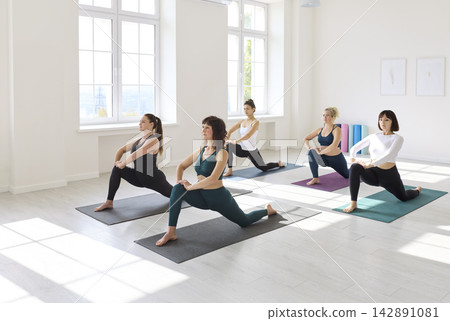 Group yoga class holding low lunge on mats in bright minimalist studio interior Group yoga class holding low lunge on mats in bright minimalist studio interior 142891081