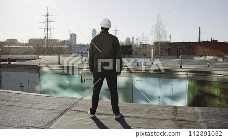 Rear view construction inspector standing on flat industrial rooftop, wearing white hard hat and olive jacket, overlooking urban skyline with concrete surfaces, metal structures, muted daylight Rear view construction inspector standing on flat industrial rooftop, wearing white hard hat and olive jacket, overlooking urban skyline with concrete surfaces, metal structures, muted daylight 142891082