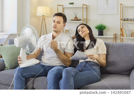Young couple sitting on sofa in living room at home and suffering from summer heat in front of fan. Young couple sitting on sofa in living room at home and suffering from summer heat in front of fan. 142891087