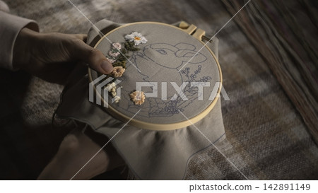 An Upclose View Of Delicate Hand Embroidery Featuring Floral Bunny On Plaid Background With Soft Warm Lighting. Media An Upclose View Of Delicate Hand Embroidery Featuring Floral Bunny On Plaid Background With Soft Warm Lighting. Media 142891149