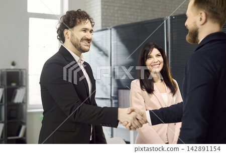 Two businessmen shake hands as smiling colleague watches in bright modern office Two businessmen shake hands as smiling colleague watches in bright modern office 142891154