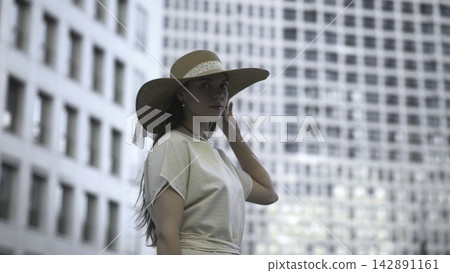 Woman in a wide hat standing before blurred city buildings Woman in a wide hat standing before blurred city buildings 142891161