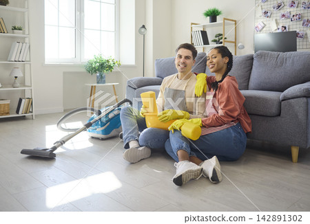 Happy couple taking break from cleaning near sofa with vacuum and supplies Happy couple taking break from cleaning near sofa with vacuum and supplies 142891302