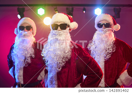 Three Santas dancing at Christmas party under colorful stage lights in shades Three Santas dancing at Christmas party under colorful stage lights in shades 142891402