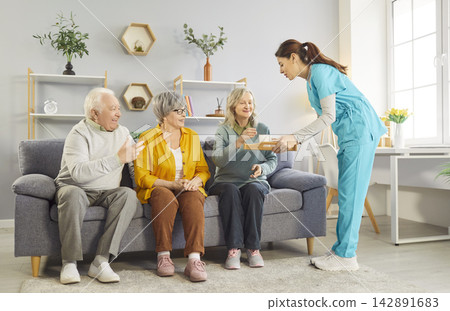 Friendly young nurse serving tea and snacks to cheerful seniors sitting on sofa in retirement home. 142891683