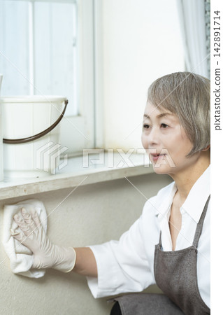 一位老婦人正在擦拭客廳裡的各種表面。 一位老婦人正在擦拭客廳裡的各種表面。 142891714