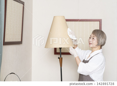 一位老婦人正在擦拭客廳裡的各種表面。 142891728