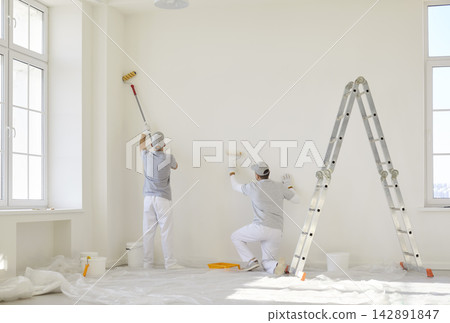 Team of professional painters in uniform painting white wall in empty room with paint rollers. Team of professional painters in uniform painting white wall in empty room with paint rollers. 142891847