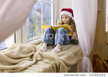 Girl in Santa hat reading book on windowsill by frosty window at Christmas Girl in Santa hat reading book on windowsill by frosty window at Christmas 142891854