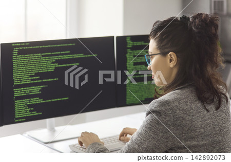Female programmer coding on dual computer screens in modern office workspace Female programmer coding on dual computer screens in modern office workspace 142892073