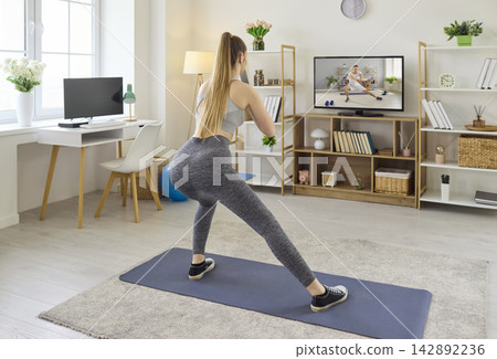 Woman doing home workout following online training on TV, stretching on yoga mat 142892236