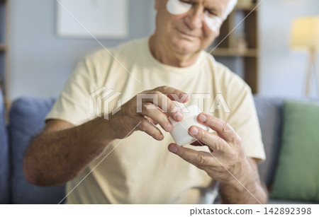 Close up of senior man with eye patches opening face cream jar at home skincare Close up of senior man with eye patches opening face cream jar at home skincare 142892398