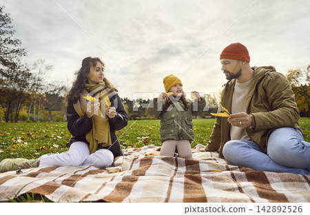 Parents and daughter playing with leaves during autumn picnic on park blanket Parents and daughter playing with leaves during autumn picnic on park blanket 142892526