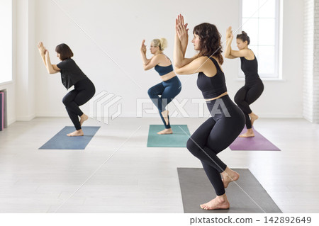 Side view of women practicing eagle pose in a minimalist yoga studio during class Side view of women practicing eagle pose in a minimalist yoga studio during class 142892649
