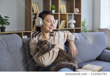 Woman relaxing at home with cat, wearing headphones and enjoying music on sofa 142892761