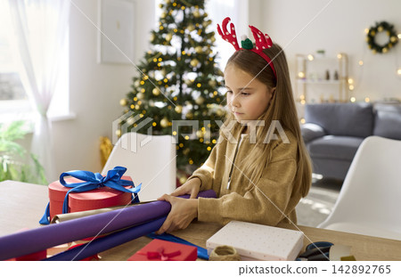 Christmas child wrapping gift with paper roll, standing at table near Xmas tree Christmas child wrapping gift with paper roll, standing at table near Xmas tree 142892765
