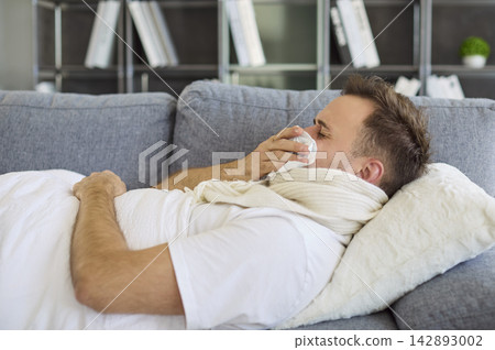 Sick man with fever sneezing into tissue while resting on sofa at home indoors 142893002
