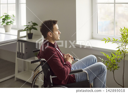 Pensive young man in wheelchair looking out window, reflecting on challenges during recovery. Pensive young man in wheelchair looking out window, reflecting on challenges during recovery. 142893018
