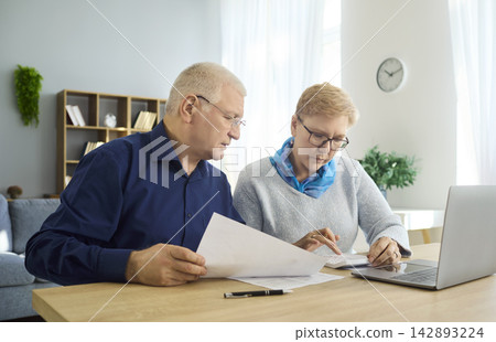 Elderly couple managing finances at home with laptop, review bills and paperwork Elderly couple managing finances at home with laptop, review bills and paperwork 142893224