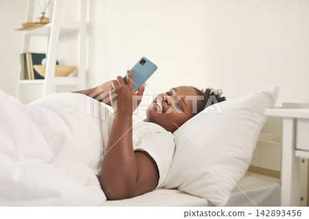 Happy African American Woman Enjoying Leisure On Her Smartphone In Bed At Home Happy African American Woman Enjoying Leisure On Her Smartphone In Bed At Home 142893456