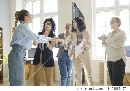 Smiling businesswomen handshake with applause at team meeting in bright office Smiling businesswomen handshake with applause at team meeting in bright office 142893471