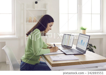 Female accountant analyzing financial data at laptops in bright home office Female accountant analyzing financial data at laptops in bright home office 142893551