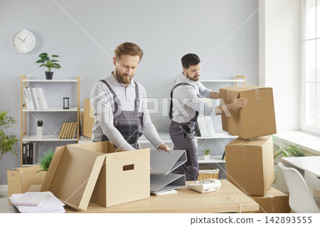 Moving office service, two men in overalls packing boxes, carrying items, placing belongings on desk Moving office service, two men in overalls packing boxes, carrying items, placing belongings on desk 142893555