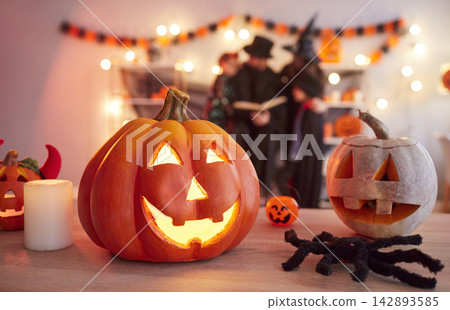 Glowing carved pumpkins standing on table while family in Halloween costumes reading book together. Glowing carved pumpkins standing on table while family in Halloween costumes reading book together. 142893585