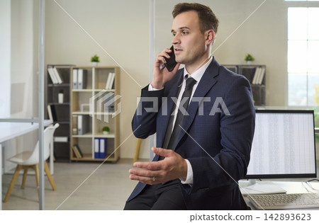 Confident businessman in suit talking on smartphone during office phone call Confident businessman in suit talking on smartphone during office phone call 142893623