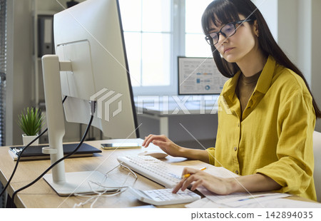 Focused woman doing finance budgeting at computer with calculator in modern office Focused woman doing finance budgeting at computer with calculator in modern office 142894035