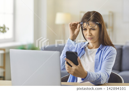 Woman with myopia, vision loss holding glasses to read text on phone screen Woman with myopia, vision loss holding glasses to read text on phone screen 142894101