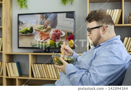 Overweight man eating salad at home while watching TV cooking show in living room Overweight man eating salad at home while watching TV cooking show in living room 142894164