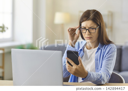 Woman with glasses squinting at phone screen, sitting at home desk with laptop Woman with glasses squinting at phone screen, sitting at home desk with laptop 142894417