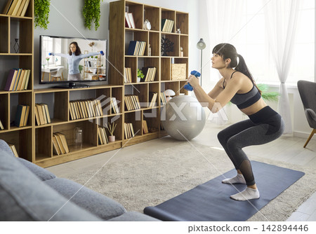 Woman doing dumbbell squats at home, online fitness class on TV in living room Woman doing dumbbell squats at home, online fitness class on TV in living room 142894446