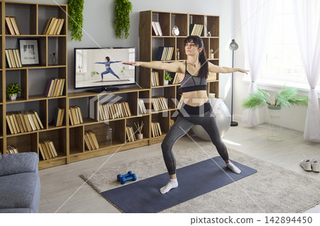 Woman doing online yoga workout at home, warrior pose training in living room Woman doing online yoga workout at home, warrior pose training in living room 142894450
