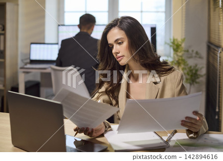 Focused businesswoman reviewing documents and working on laptop at modern office Focused businesswoman reviewing documents and working on laptop at modern office 142894616