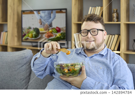 Overweight man eating salad at home while watching a TV cooking show on sofa 142894784