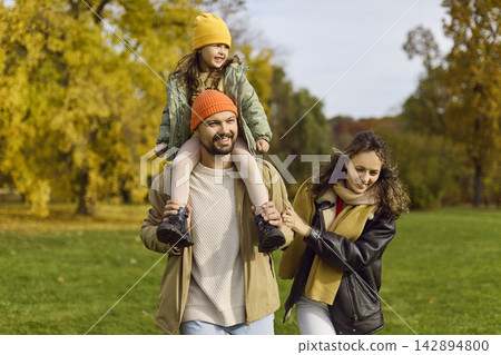 Father carrying daughter as parents walk through vibrant autumn park together Father carrying daughter as parents walk through vibrant autumn park together 142894800
