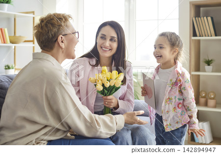 Child girl with mother congratulating grandmother on holiday sitting on sofa at home. Child girl with mother congratulating grandmother on holiday sitting on sofa at home. 142894947