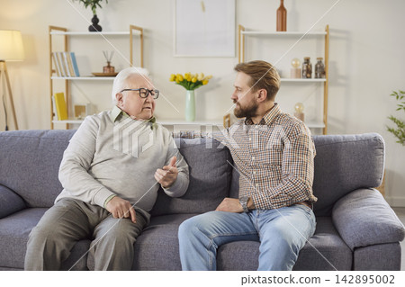 Serious senior father talking with adult son while sitting on sofa and sharing thoughts at home. Serious senior father talking with adult son while sitting on sofa and sharing thoughts at home. 142895002