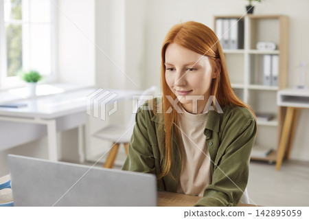 Portrait of a young woman sitting at the desk on her workplace w and working on a laptop. Portrait of a young woman sitting at the desk on her workplace w and working on a laptop. 142895059