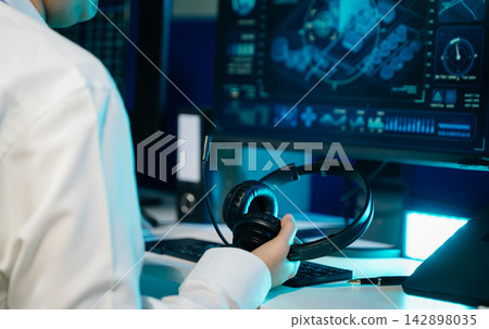 Focused professional holding a headset at a modern operations workstation, monitoring network security data with a colleague in a high tech control room environment. Focused professional holding a headset at a modern operations workstation, monitoring network security data with a colleague in a high tech control room environment. 142898035