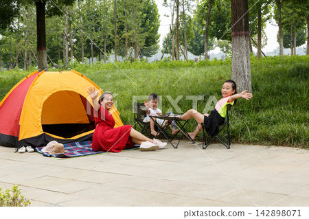 Young family enjoying summer camping in city park with orange tent under green trees Young family enjoying summer camping in city park with orange tent under green trees 142898071