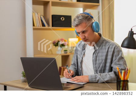 Middle-aged man on home table learning from online course on laptop pausing video and writing notes Middle-aged man on home table learning from online course on laptop pausing video and writing notes 142898352